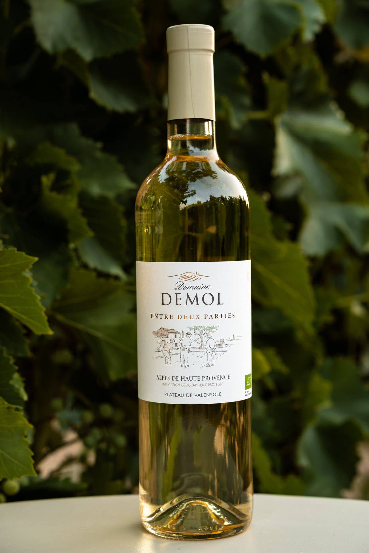 Bouteille de vin blanc Entre deux Parties du Domaine Demol, étiquette de face, devant un feuillage de vigne