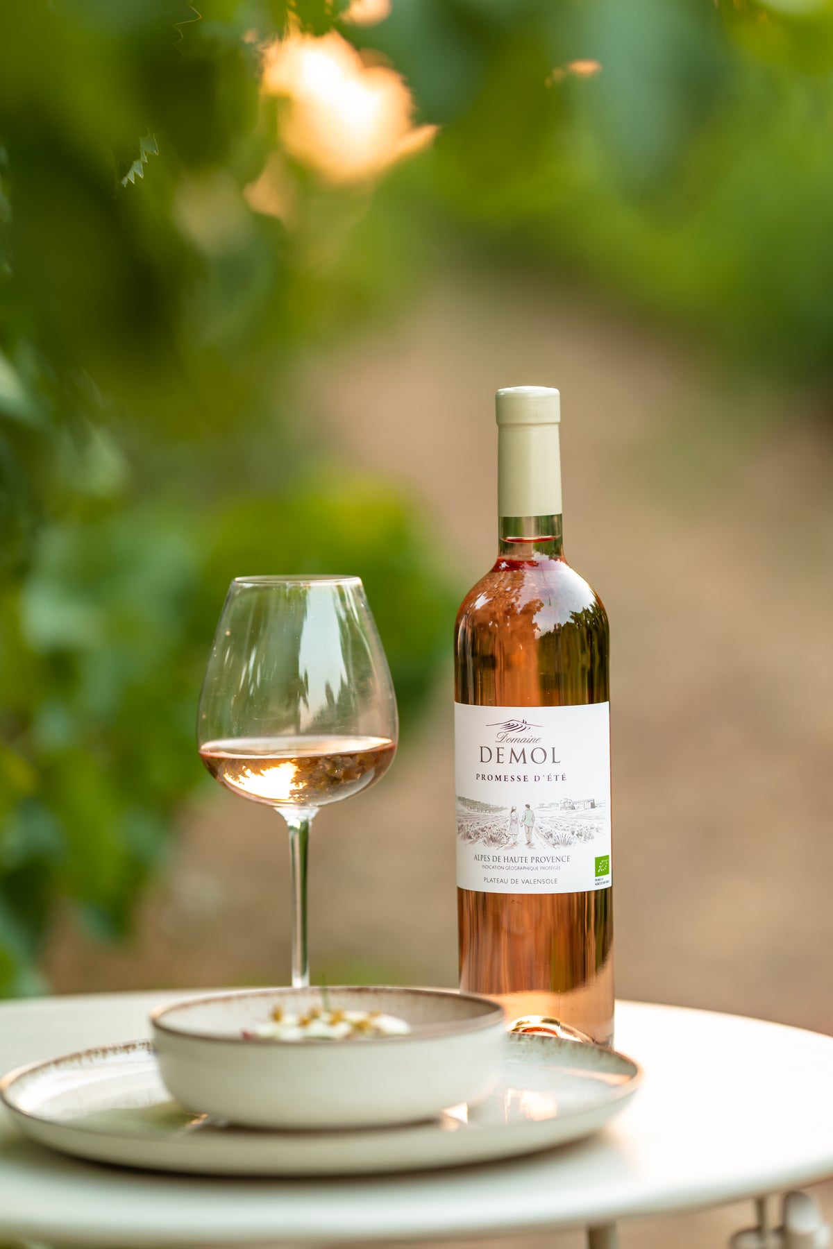 Bouteille de rosé Promesse d'été du Domaine Demol, un verre servi et une assiette, au milieu des vignes
