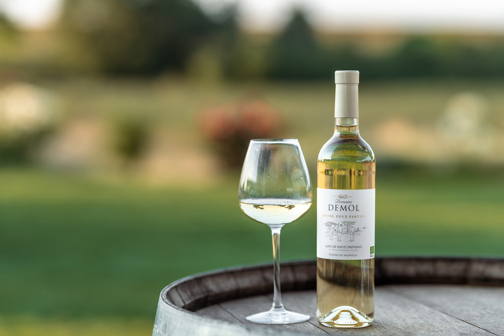 Bouteille de vin blanc Entre deux Parties du Domaine Demol et un verre servi, posés sur un tonneau