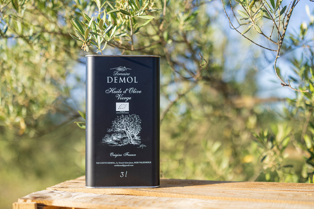 Bidon de 3 l d'huile d'olive vierge Domaine Demol, étiquette noire, sur une table en bois sous un olivier